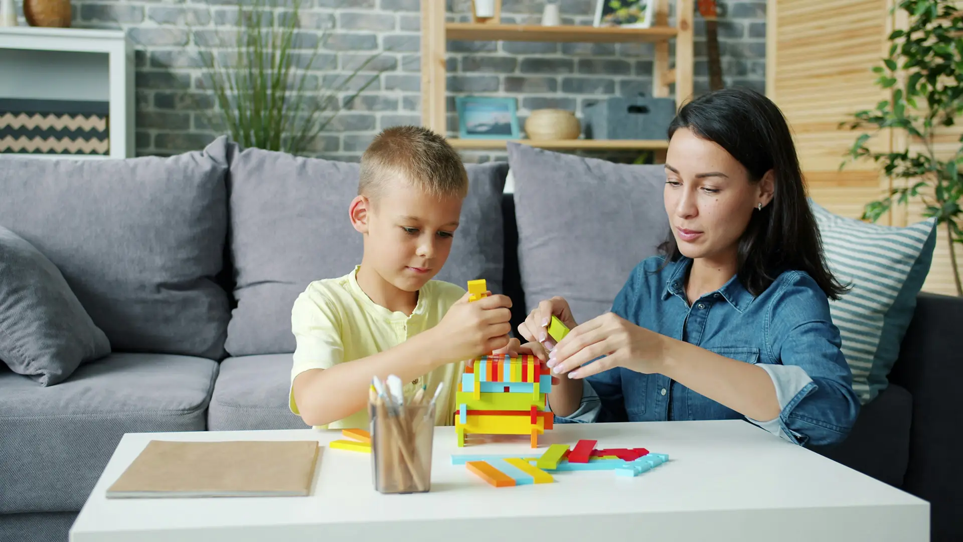 Woman and boy sitting at table building structure with colorful interlocking blocks.