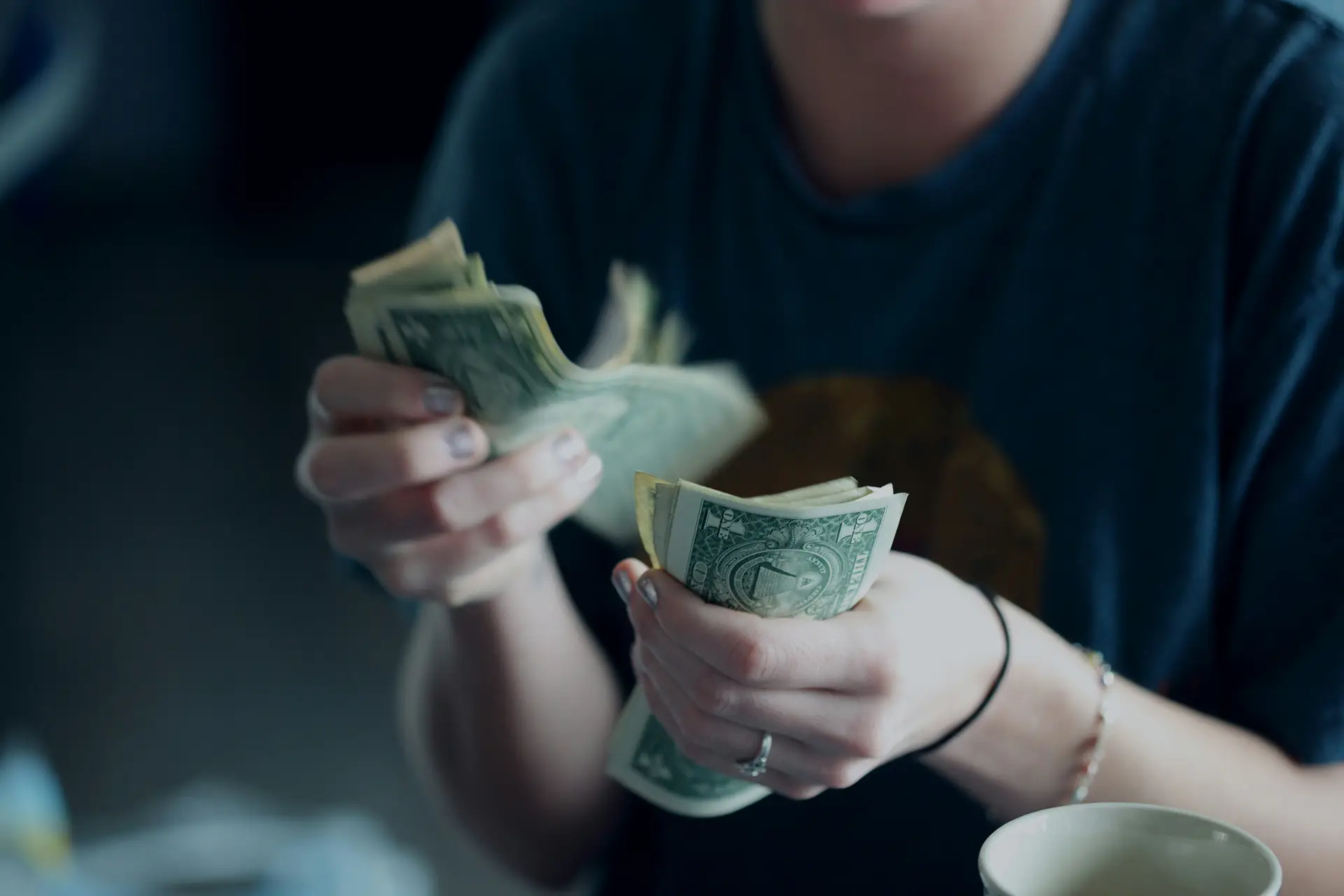 Person holding and counting a stack of U.S. dollar bills with a coffee cup nearby.