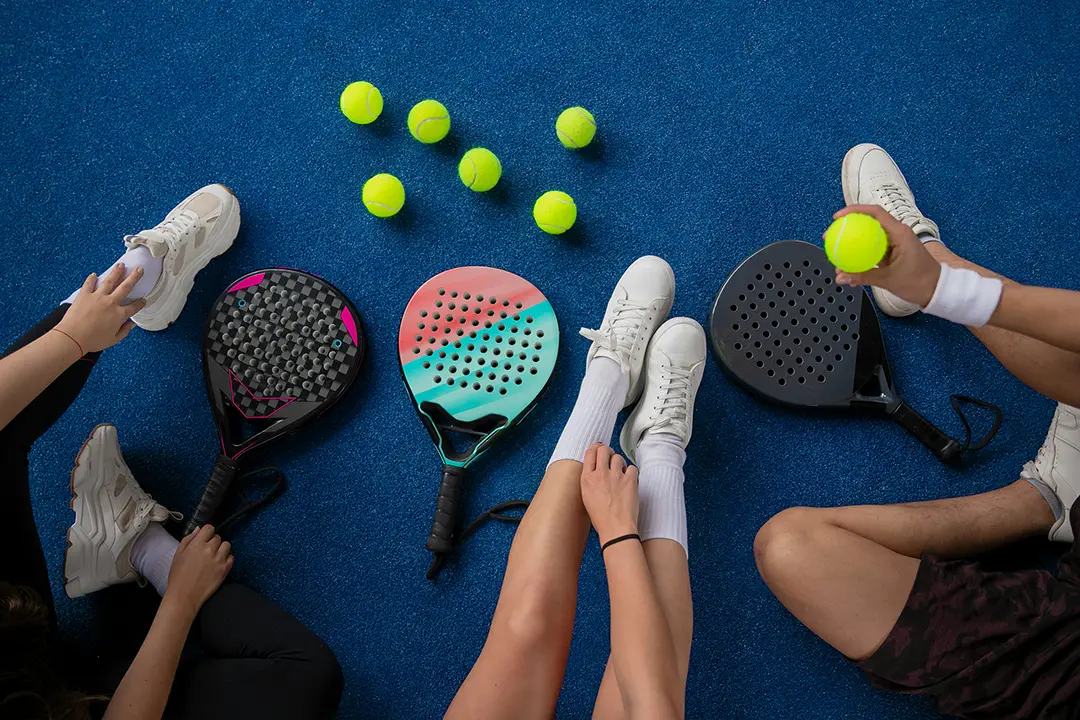 Three people sitting on blue court with three padel rackets and seven yellow padel balls.