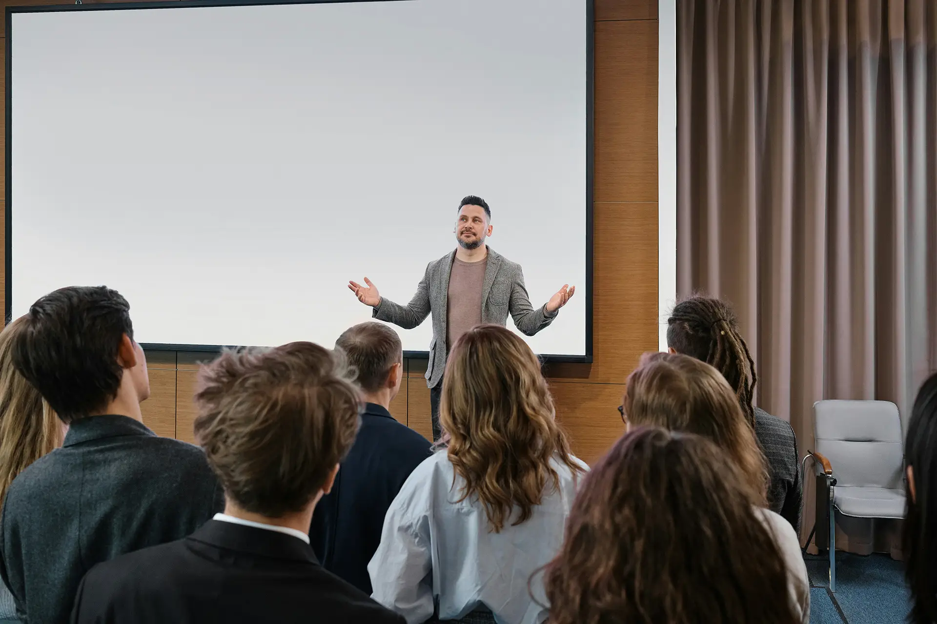 Man in a blazer speaking to an audience in a conference room with a blank projector screen.