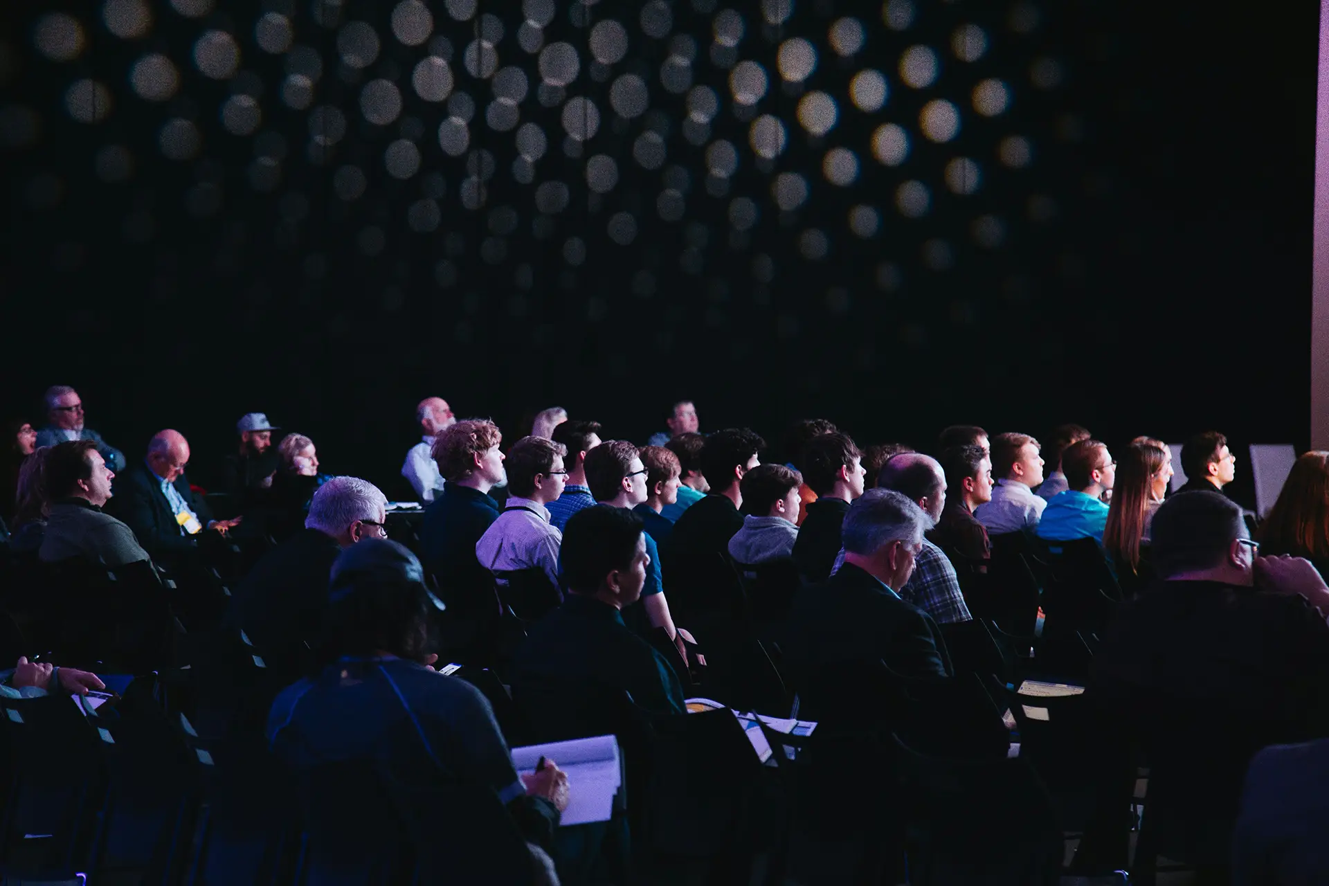Audience seated in a dark room attentively watching an unseen speaker or presentation.
