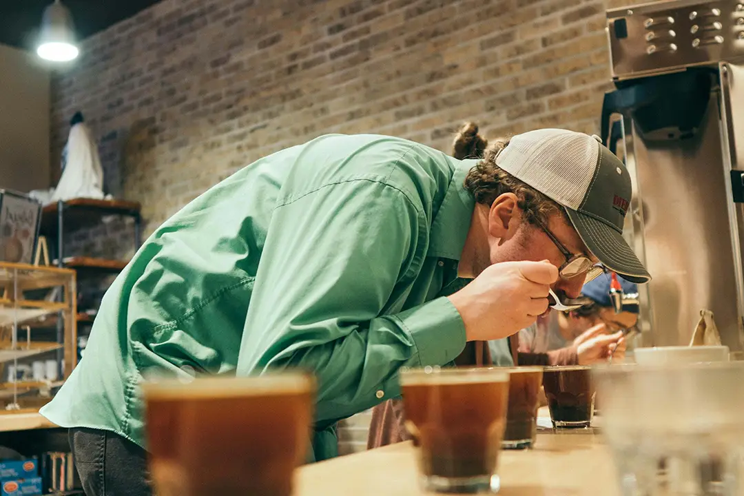 Man in green shirt and cap smelling coffee from a spoon at a coffee tasting event.