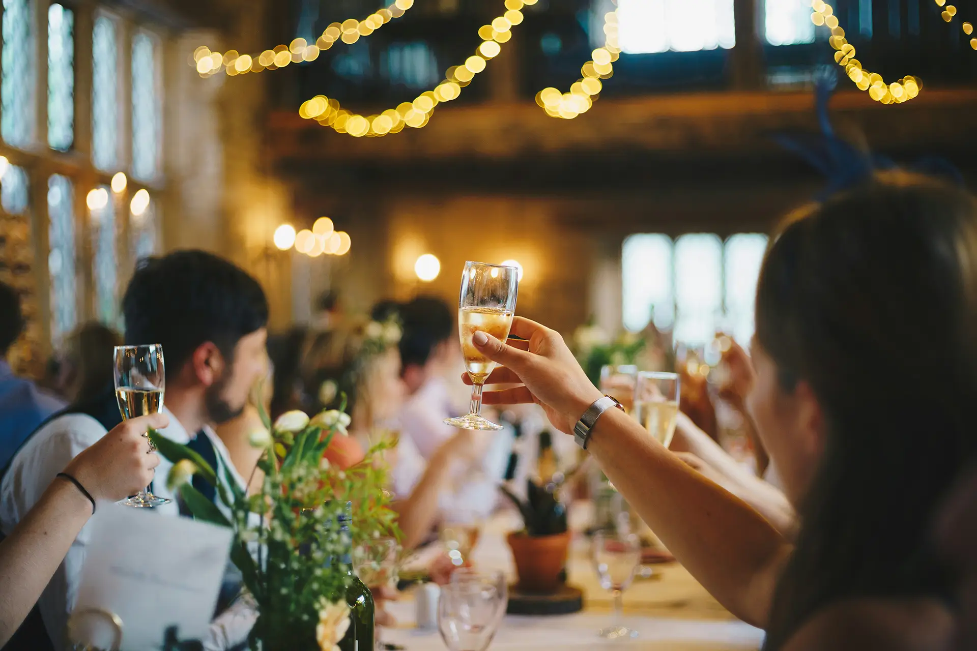 People raising champagne glasses in a warm, decorated room with string lights and blurred background.