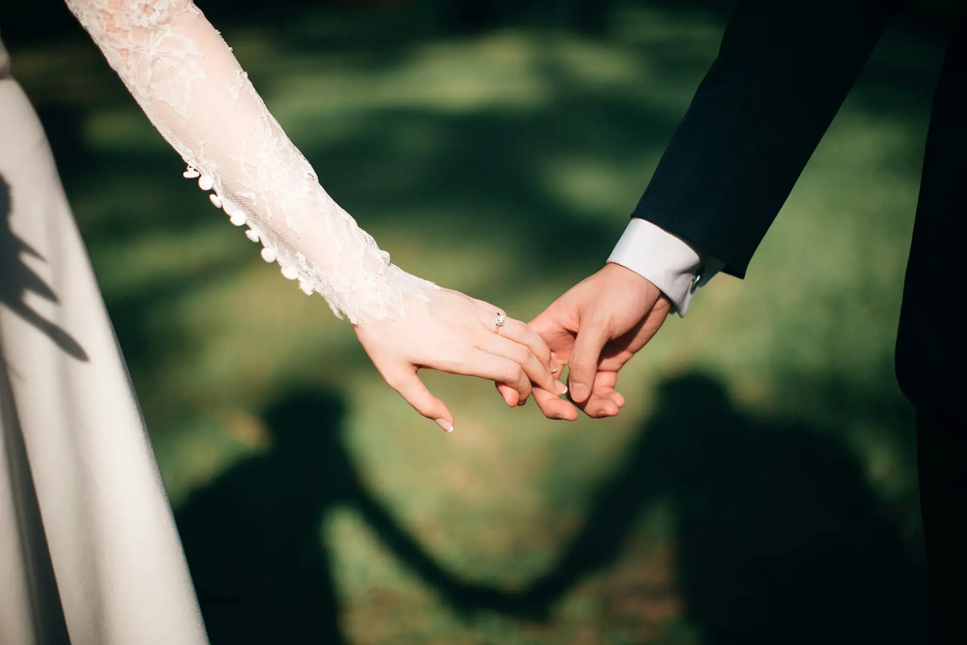 Bride and groom holding hands outdoors showing wedding ring and shadows on grass.